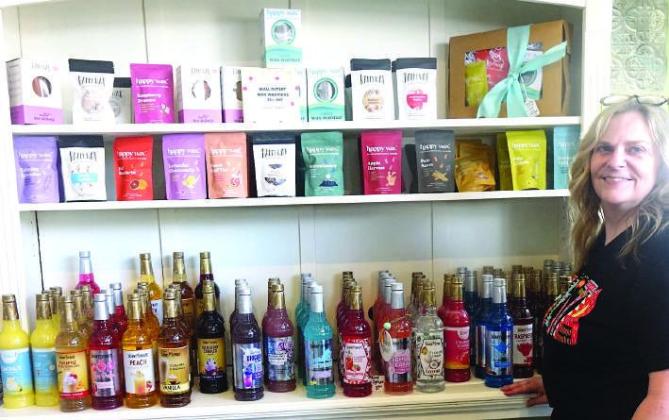 Jana O’Rear stands by a wall section of her mercantile store which is full of things to. sample. The bag items at top are Happy Wax Warmers which will melt wax to give relaxing scents.The bottles below are of the many varieties of Jordan’s Skinny Syru “Sample Saturday”
